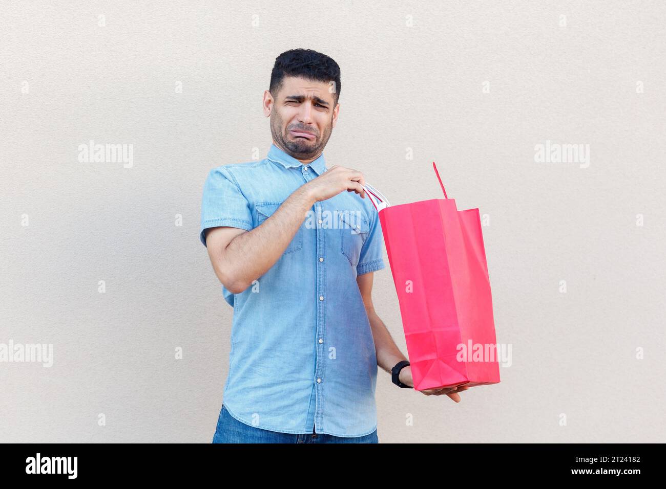 Porträt eines traurigen, gestressten, depressiven Mannes, der ein Jeanshemd trägt, enttäuscht von seinem Einkaufen im Einkaufszentrum ist, Papiertüte hält und weint. Innenstudio isoliert auf grauem Hintergrund. Stockfoto