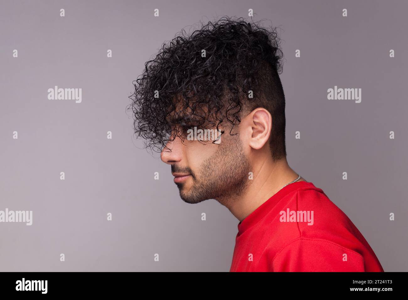 Seitliches Porträt eines attraktiven, hübschen jungen, bärtigen Mannes mit lockigen Haaren, der einen roten Pullover trägt, nach vorne blickt und seine Frisur zeigt. Innenstudio isoliert auf grauem Hintergrund. Stockfoto
