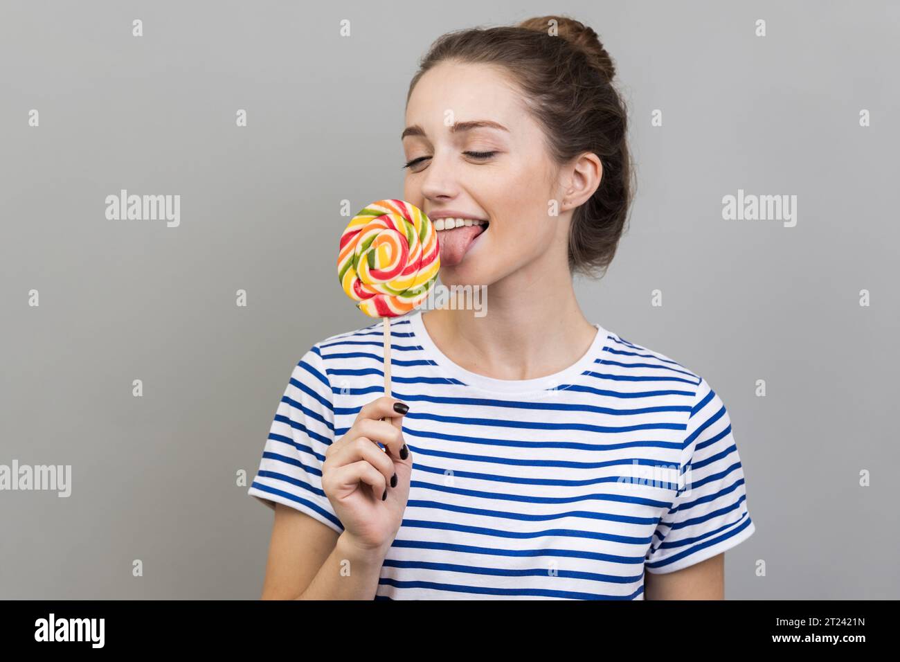Porträt einer glücklichen Frau, die ein gestreiftes T-Shirt trägt und mit ausgezogener Zunge steht, träumt und versucht ist, Lutscher zu schmecken, runde süße Süßigkeiten aus dem Regenbogen. Innenstudio isoliert auf grauem Hintergrund. Stockfoto