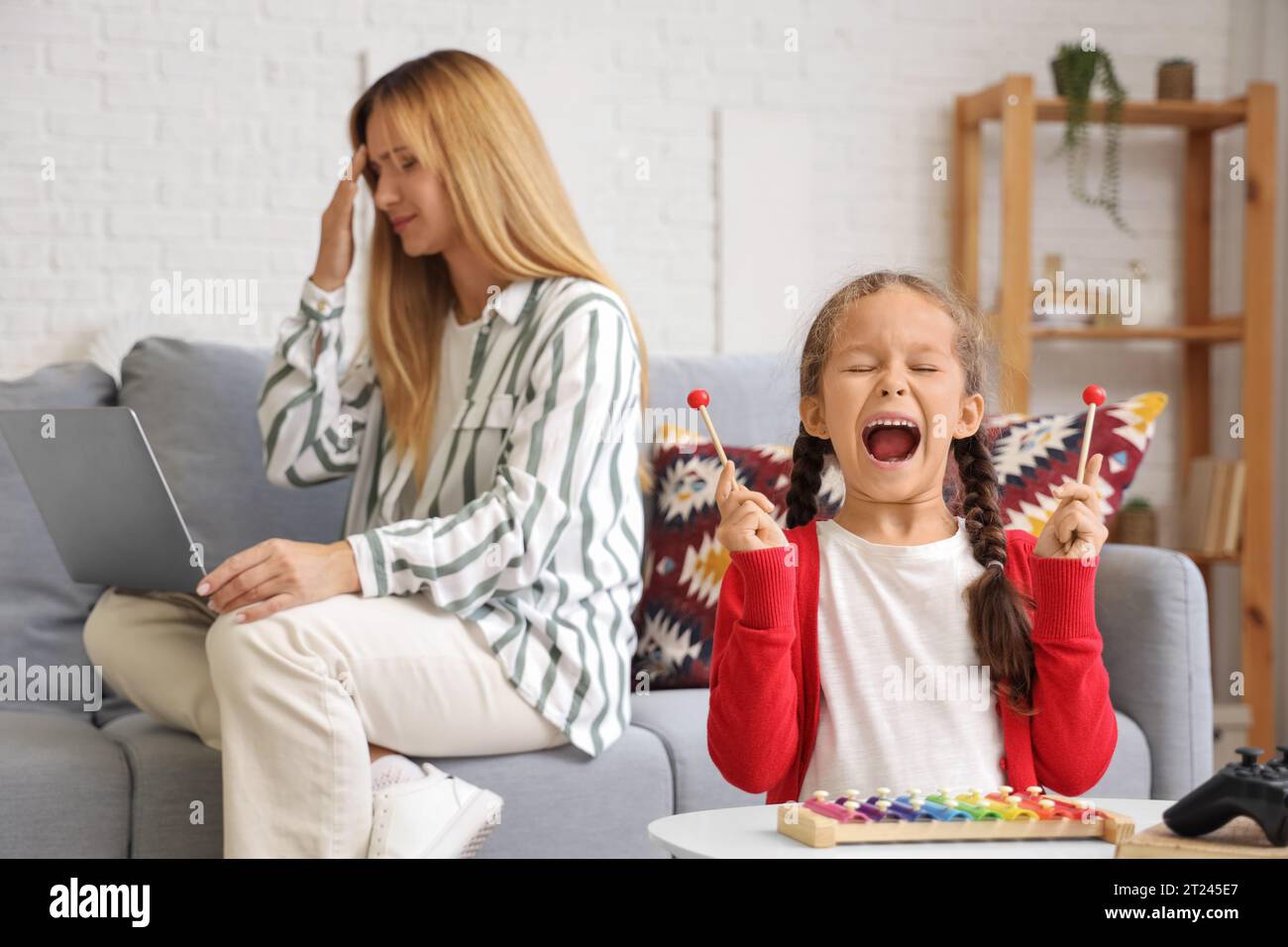 Lautes kleines Mädchen, das Xylophon spielt, und ihre arbeitende Mutter zu Hause Stockfoto