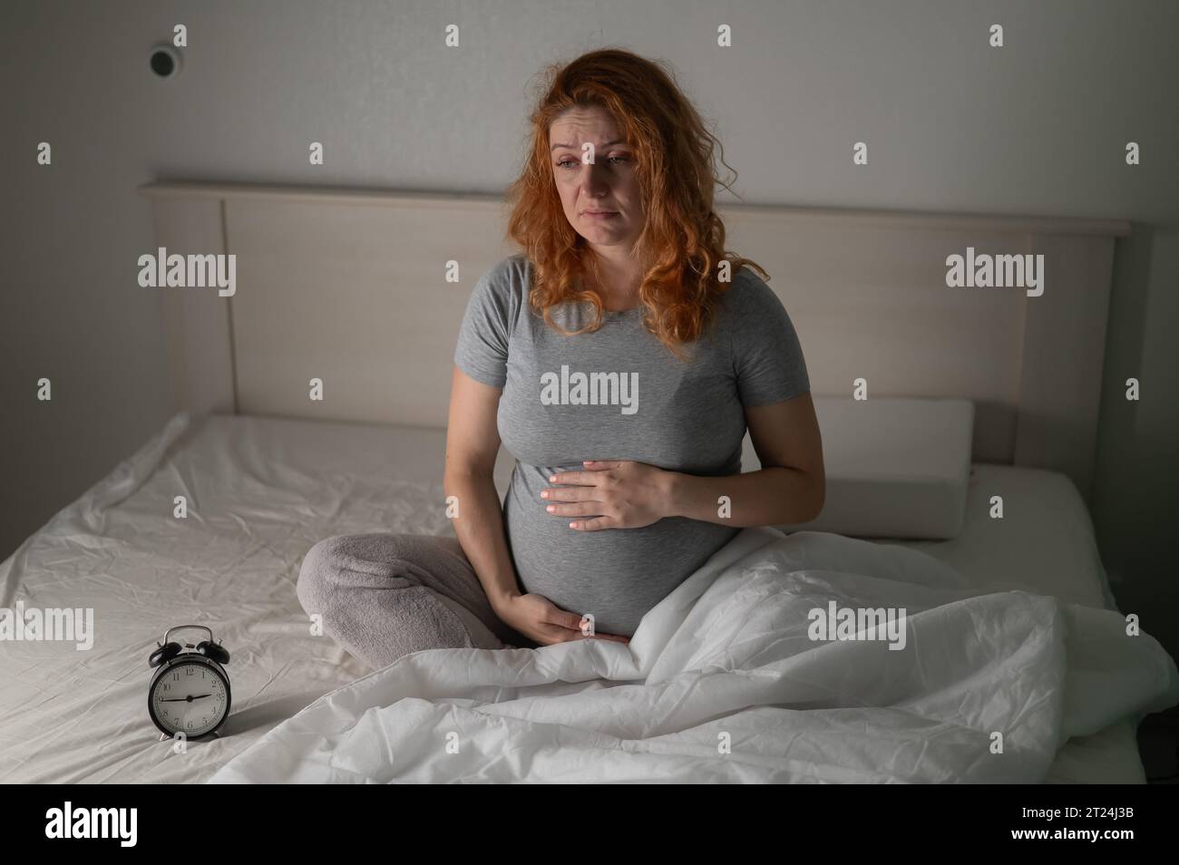 Eine schwangere Frau sitzt im Bett und schaut auf den Wecker. Schlaflosigkeit. Stockfoto