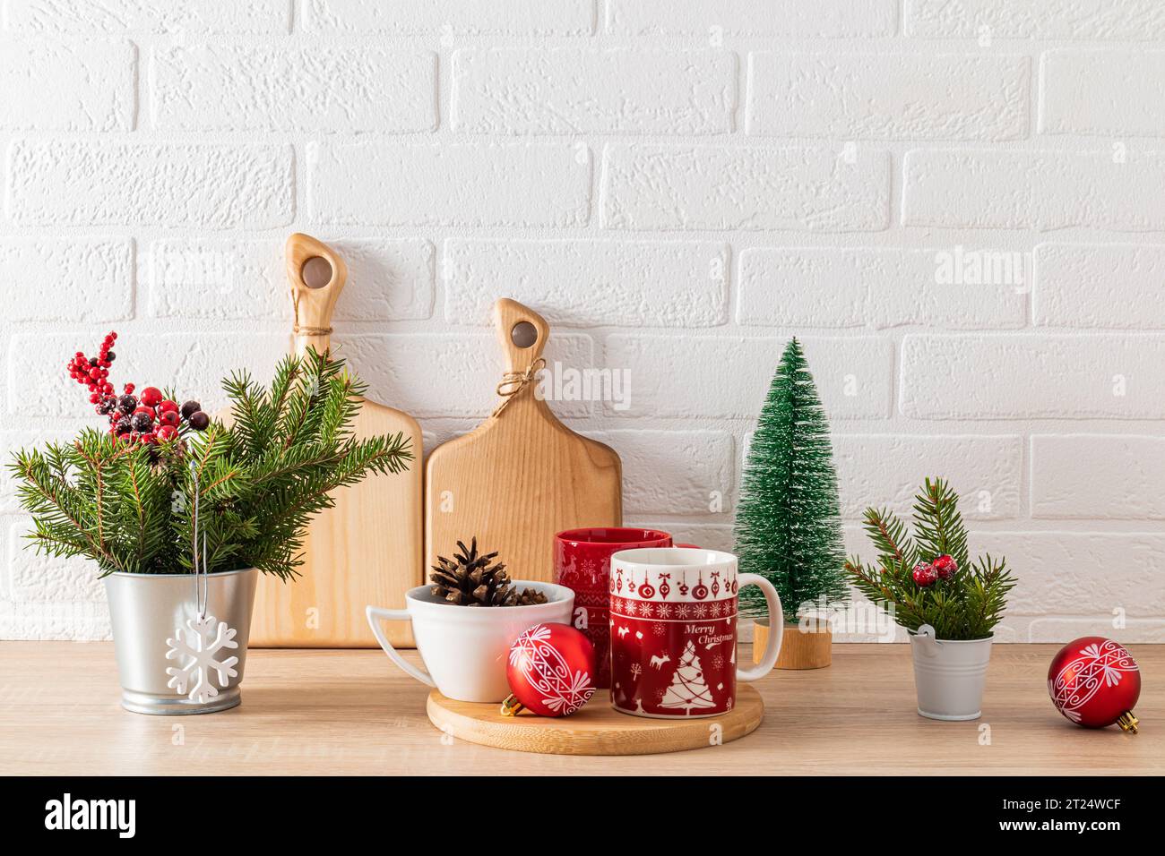 Rote Tassen mit weihnachtlichem Muster auf Holztablett zwischen grünen Dekorationen mit Tannenzweigen auf der Küchenarbeitsplatte. Gemütliches Zuhause Stockfoto