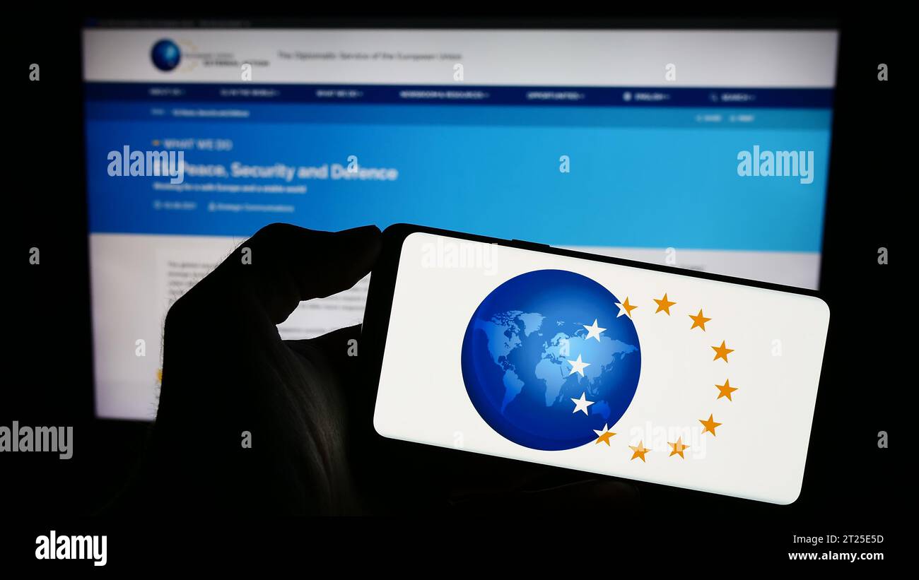 Person, die ein Mobiltelefon mit dem Logo des Europäischen Auswärtigen Dienstes (EAD) vor der Webseite hält. Schwerpunkt auf dem Display des Telefons. Stockfoto