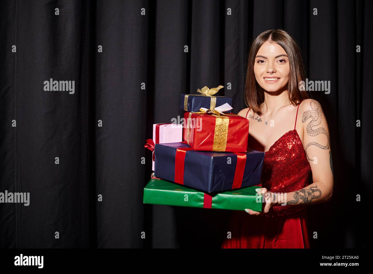 Fröhliche junge Frau, die Geschenke hält und in die Kamera lächelt, mit Vorhängen im Hintergrund, Weihnachtsgeschenke Stockfoto