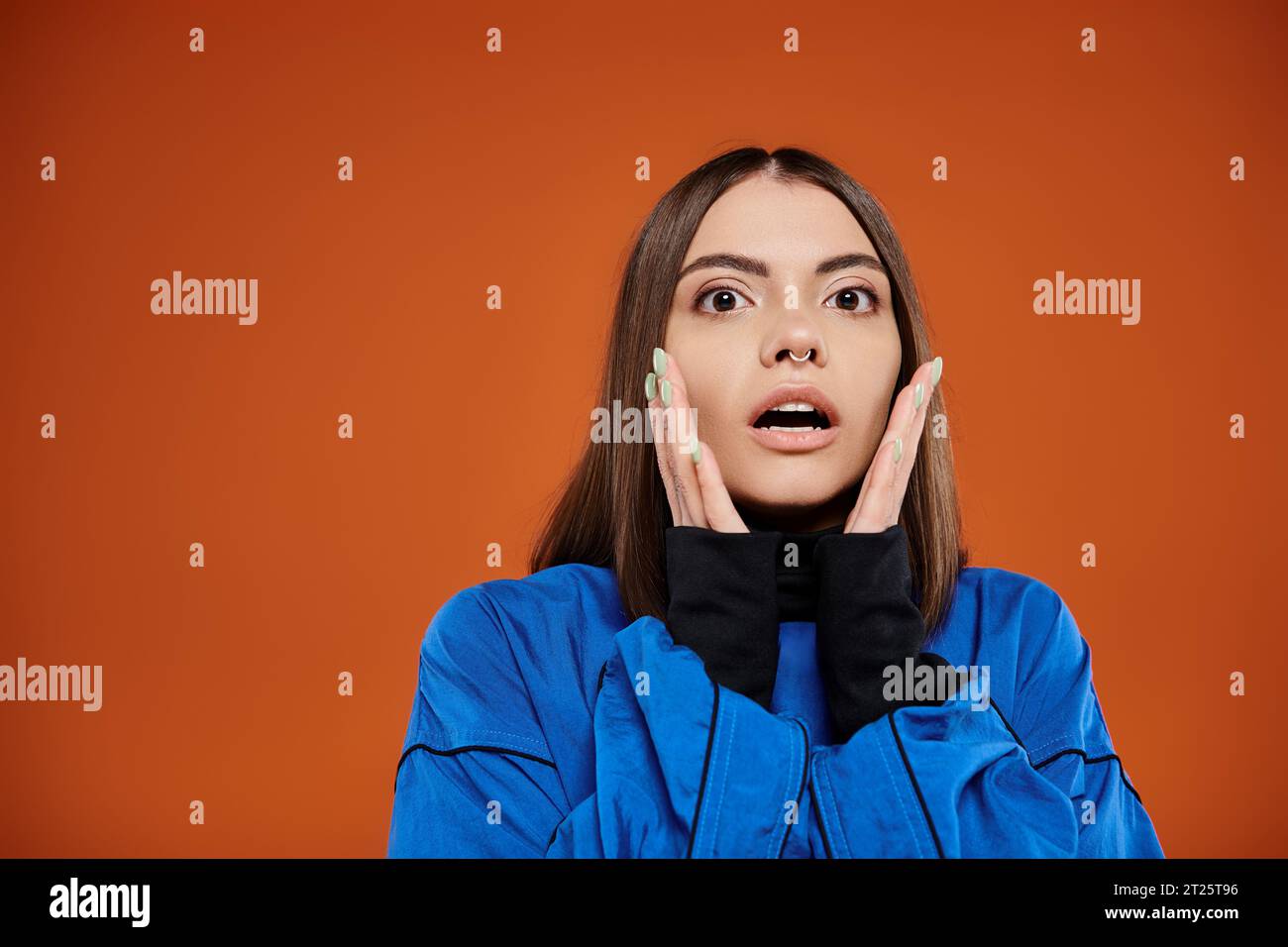 Schockierte Frau mit durchbohrter Nase, die Wangen mit den Händen berührt und die Kamera vor orangefarbenem Hintergrund betrachtet Stockfoto