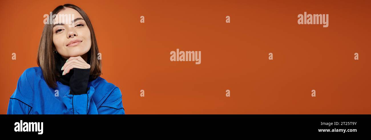 Nachdenkliche Frau mit durchbohrter Nase, die in die Kamera schaut, während sie vor orangefarbenem Hintergrund denkt, Banner Stockfoto