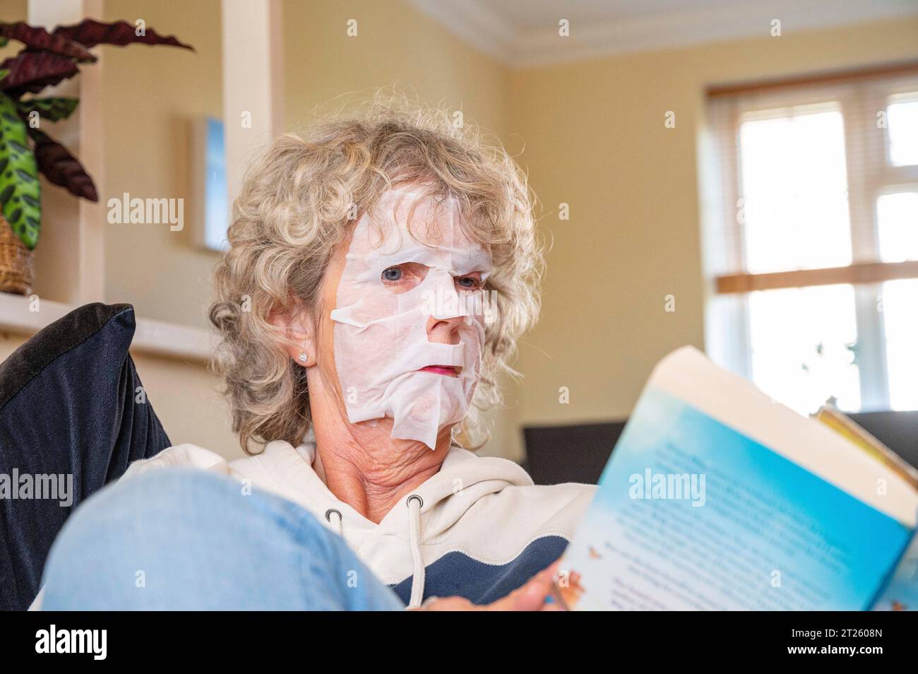 Reife Frau, die zu Hause ein Buch liest, trägt eine Gesichtspackung, um unerwünschte Falten loszuwerden und ihre Haut glatt zu halten Stockfoto