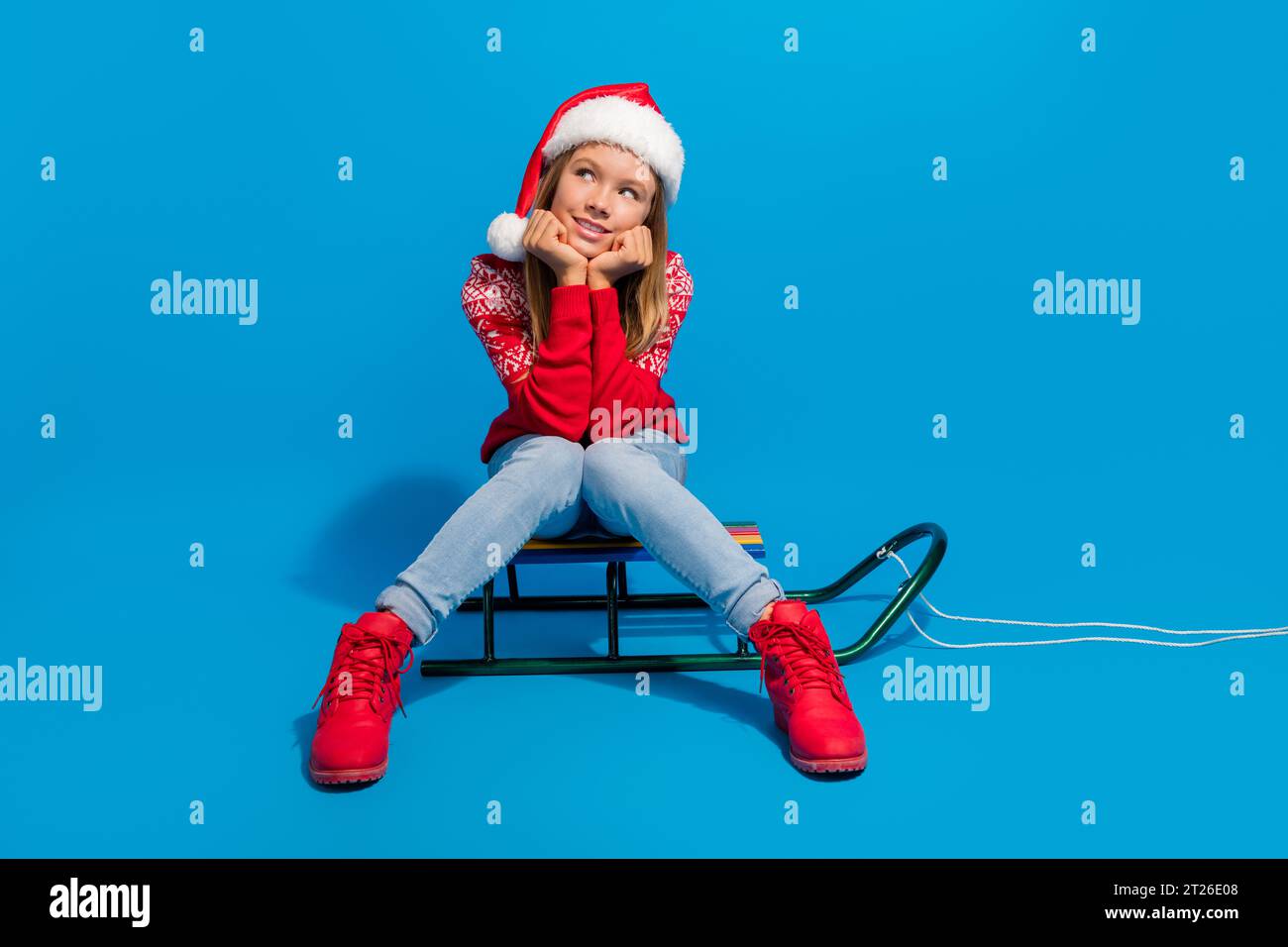 Foto in voller Länge von traumhaften Teenager Mädchen tragen Pullover santa Hut sitzen auf Schlitten Blick auf Verkauf leeren Raum isoliert auf blauem Hintergrund Stockfoto