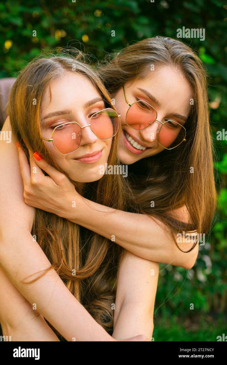 Erstaunte glückliche junge Zwillingsschwestern Generation Zkuschelte sich mit Sonnenbrille in einem Park, Frauen posierten draußen. Konzept der Freundschaft Happy Girls Umarmung Stockfoto