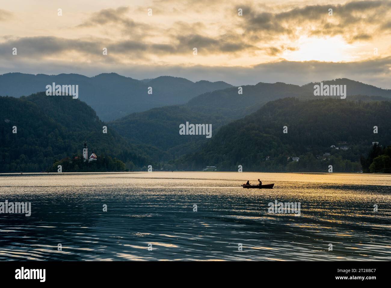 Menschen in einem Ruderboot mit der Insel Bled (dunkler, bewölkter Himmel) inmitten des Bleder Sees, slowenische Alpen, Slowenien. Stockfoto