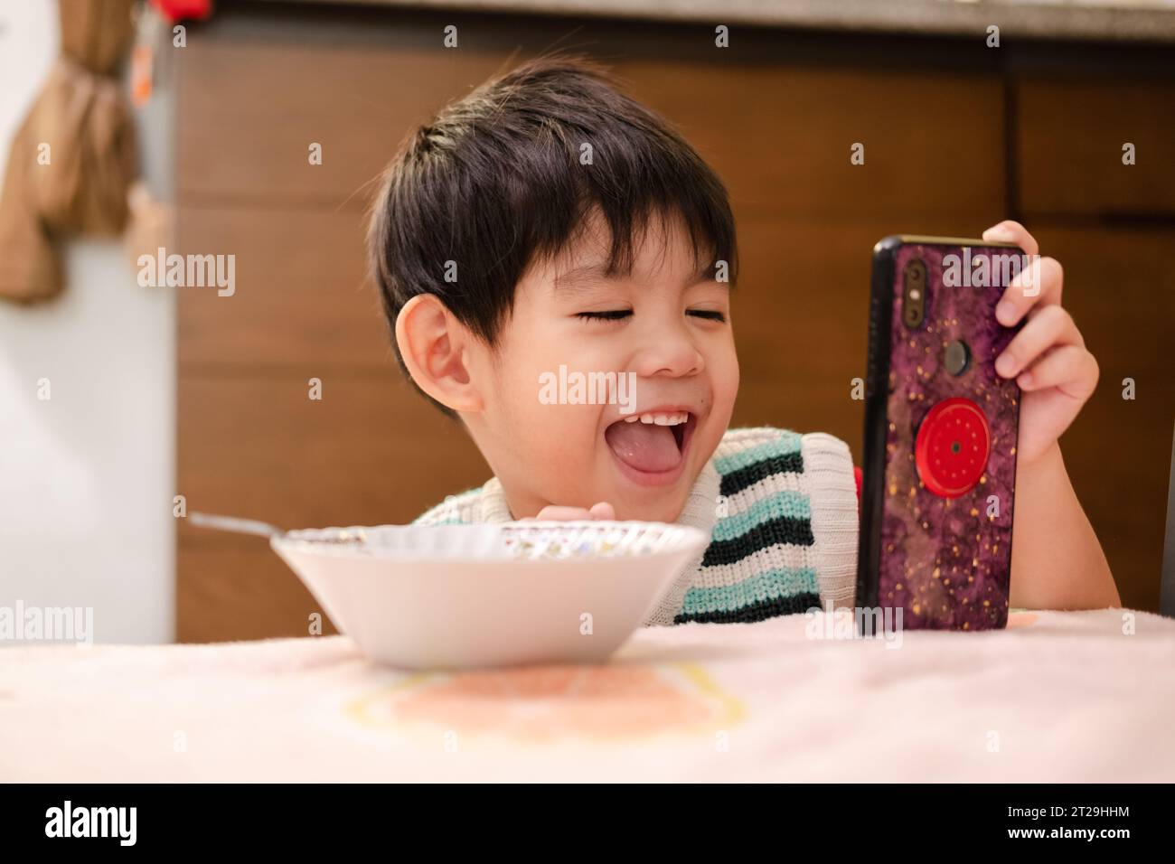 Das Gesicht eines asiatischen Kindes, das beim Anblick des Telefons isst Stockfoto