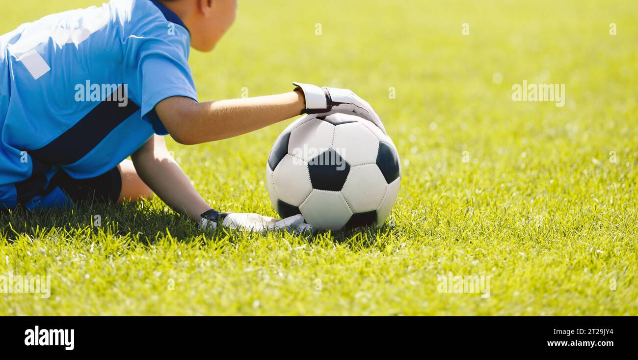 Ein kleiner Junge als Fußballtorwart, der während eines Turnierspiels einen Fußball fängt. Ein Schulfußball-Torwart in einem blauen Trikot und Sporthandschuhen Stockfoto