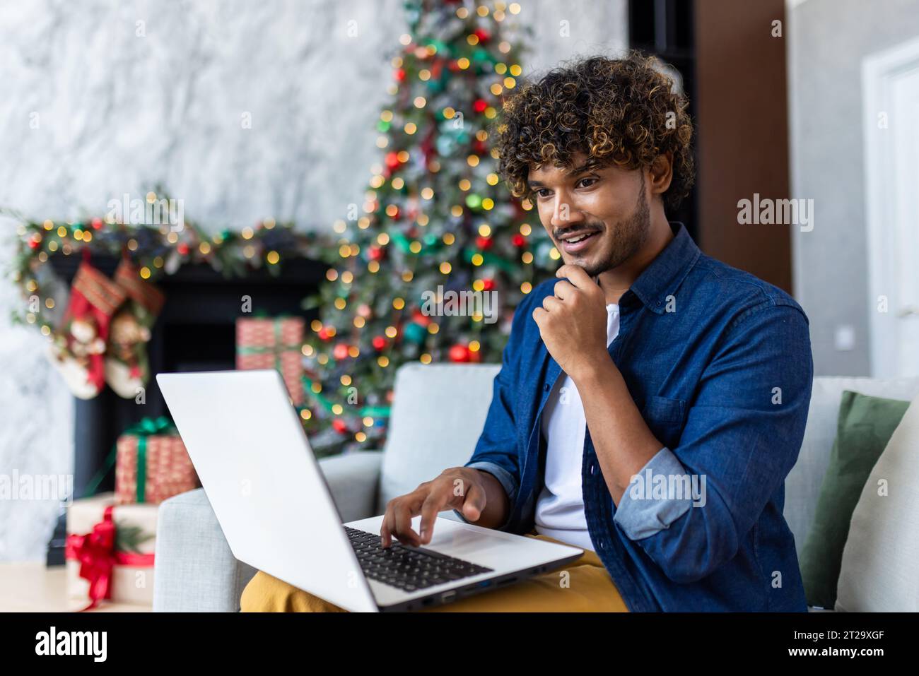 Ein junger Mann sitzt auf Sofa im Wohnzimmer an einem Wintertag zu Weihnachten. Ein lateinamerikanischer Mann sucht Waren und Geschenke online in einem Online-Shop aus, benutzt einen Laptop, in der Nähe eines geschmückten Weihnachtsbaums. Stockfoto