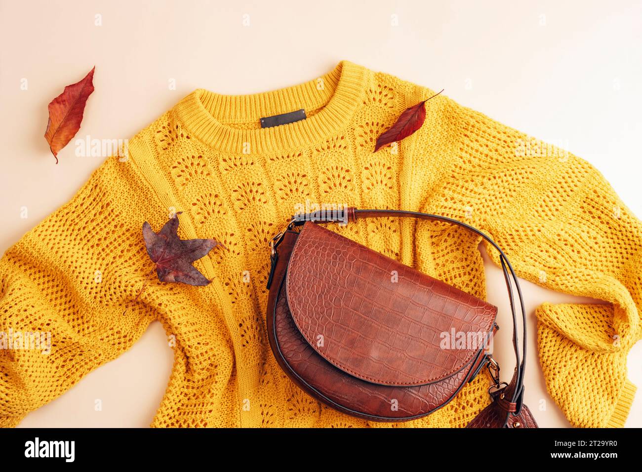 Flache Lage des gelben Pullovers und der braunen Tasche der Frau, Herbstblätter auf beigefarbenem Hintergrund. Herbstmodekonzept. Draufsicht, flach. Stockfoto