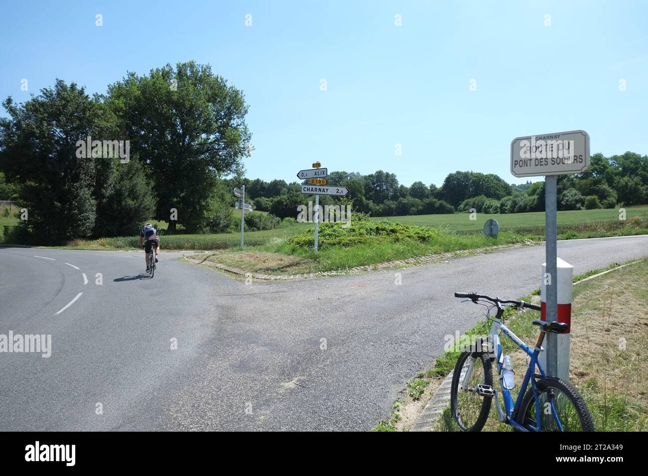 Ein Radfahrer überquert eine Abzweigung in der Straße, und ein Fahrrad beugt sich gegen ein Straßenschild, Route du Pont des Soupirs, Bäume, Straßen, ein Schild nach Charnay in Rhône Frankreich Stockfoto
