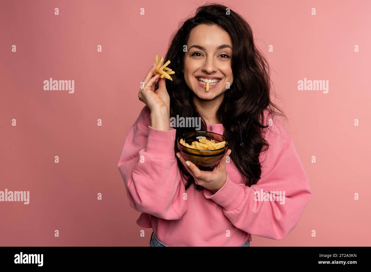 Fröhliche junge Frau isst frittierte Kartoffeln aus der Schüssel während des Fotoshootings im Studio. Konzept für den Fast Food-Konsum Stockfoto