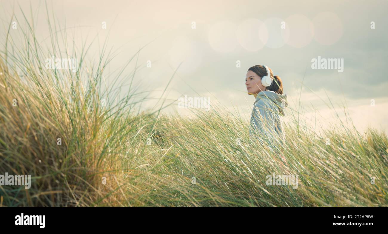 Frau mit Kopfhörern, die durch langes Gras geht und einen Hoodie trägt. Vom Konzept weg zu kommen. Stockfoto