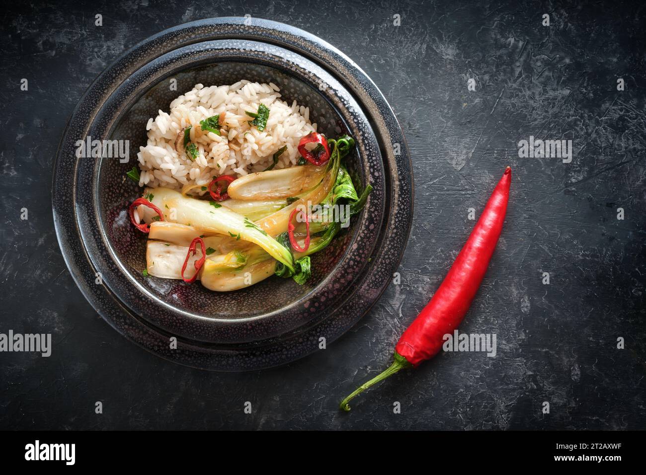 Asiatisches Gericht aus Reis ad Pak Choi (Chinakohl) und roter Chilischote in einer schwarzen Keramikschale mit Knoblauch, Ingwer und Kräutern auf dunklem Hintergrund, Co Stockfoto