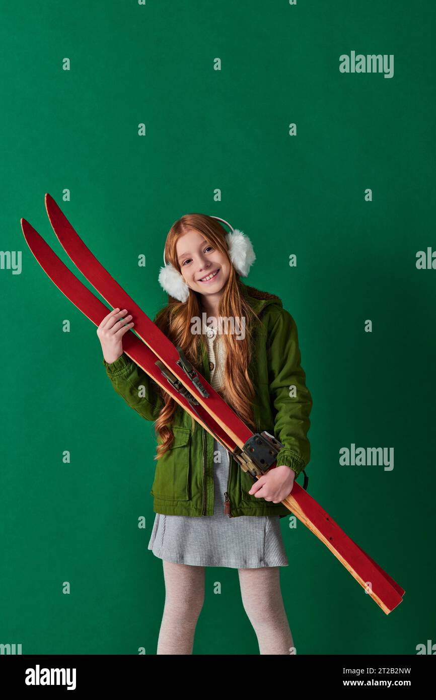 Fröhliches Mädchen in Ohrenschützern und Winteroutfit mit roter Skiausrüstung vor türkisfarbenem Hintergrund Stockfoto