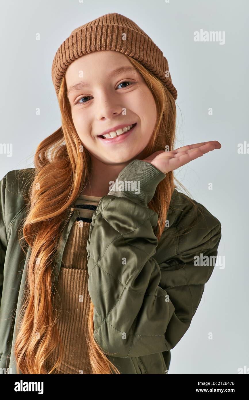 Wintermode, glückliches Mädchen mit langen Haaren und Strickmütze in khakifarbener Jacke auf grauem Hintergrund Stockfoto