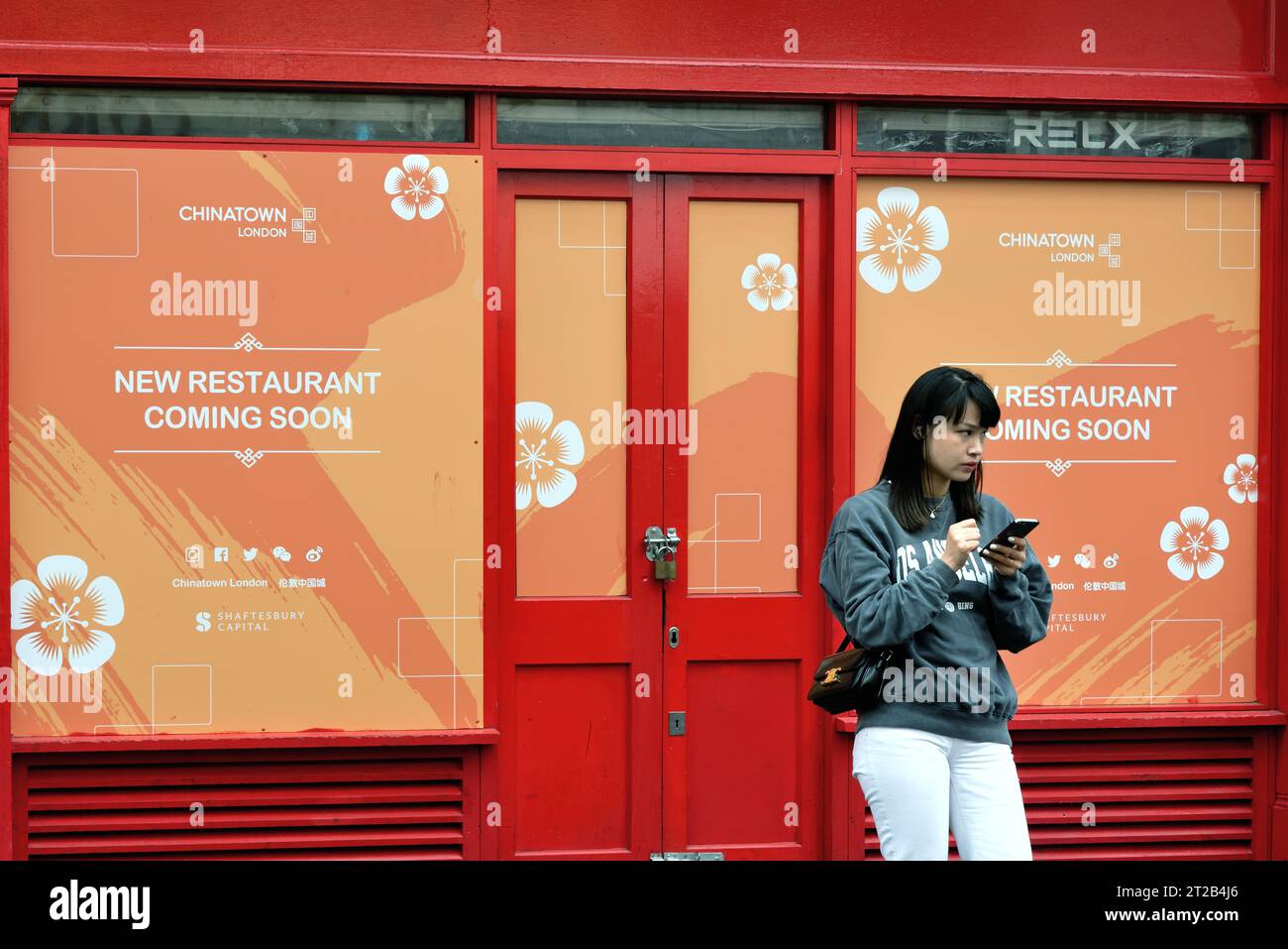 Eine junge Chinesin, die ein Mobiltelefon vor einer rot-gelben Restaurantfassade benutzt, die bald in Chinatown Wardour Street London, Großbritannien, eröffnet wird Stockfoto