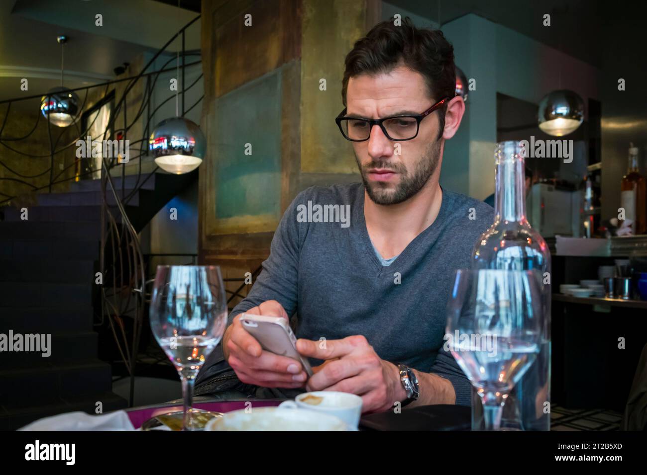 Ein hübscher Mann mit Brille schaut auf ein Handy, während er in einem Restaurant in Paris sitzt. Stockfoto