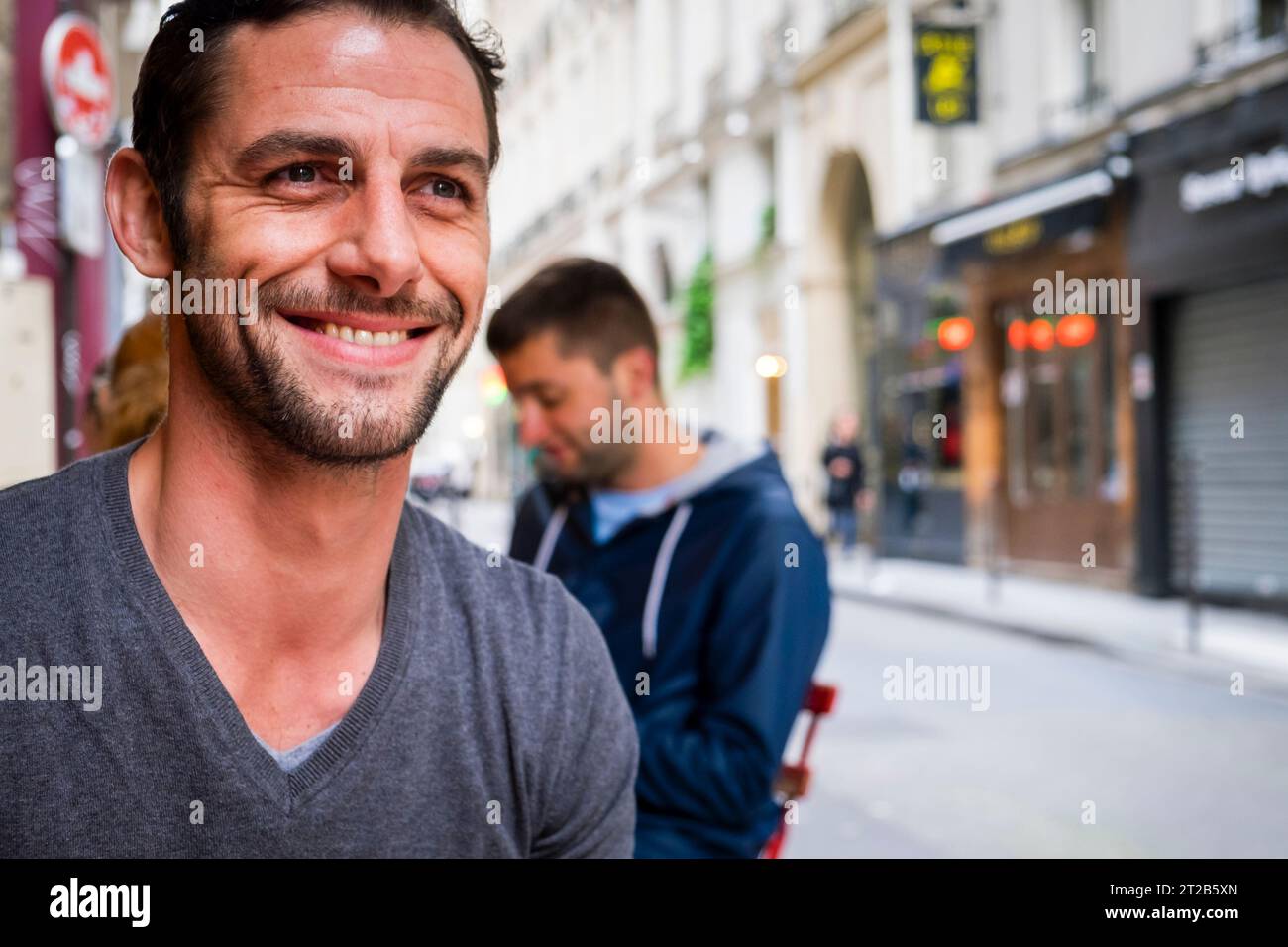 Ein hübscher Mann lächelt und schaut von der Kamera weg, während er in einem Café im Freien in Paris sitzt. Stockfoto