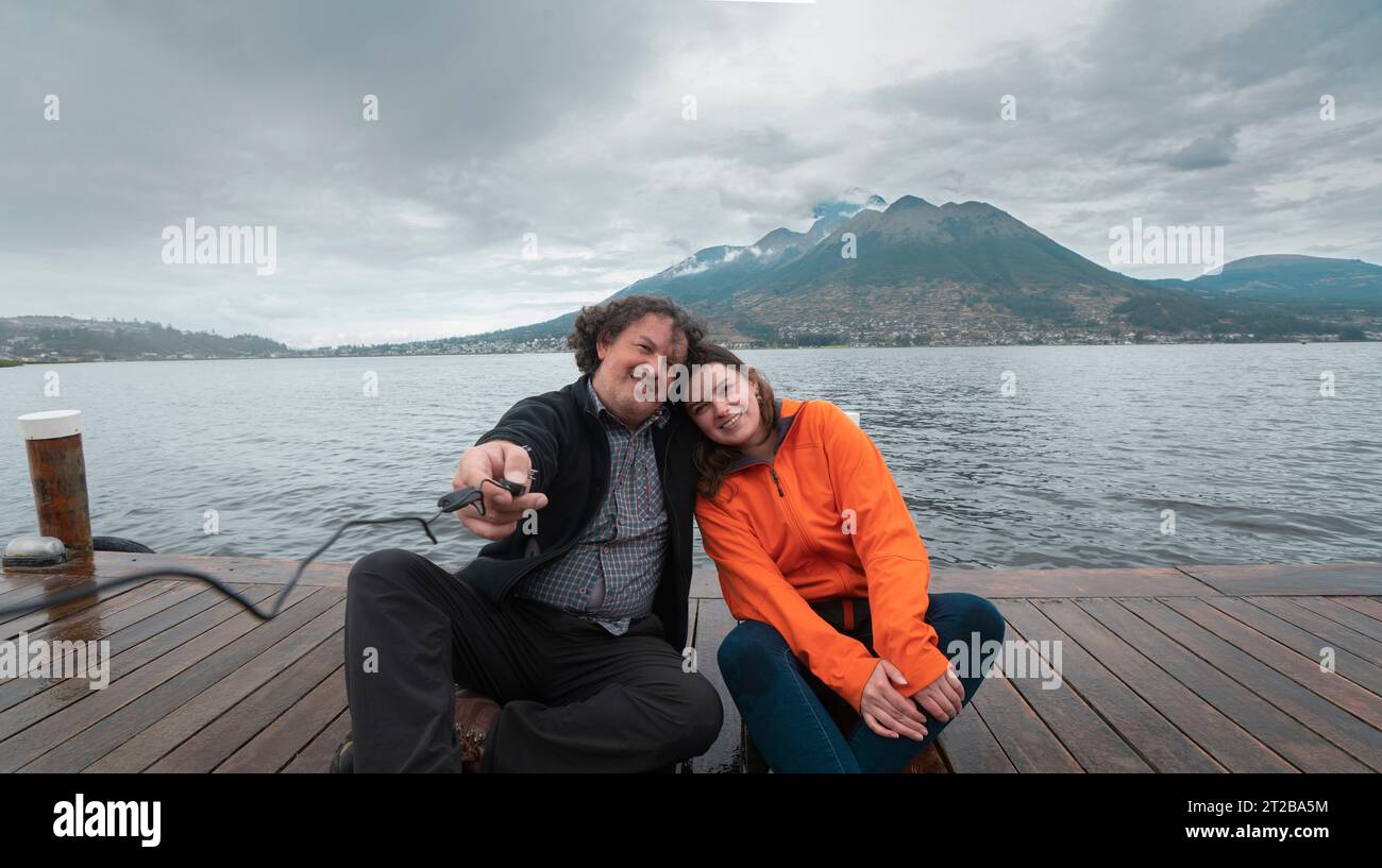 Das junge lateinamerikanische Paar sitzt auf einem hölzernen Pier am Lake San Pablo und macht an einem bewölkten Tag ein Selfie mit dem Vulkan Imbabura im Hintergrund Stockfoto