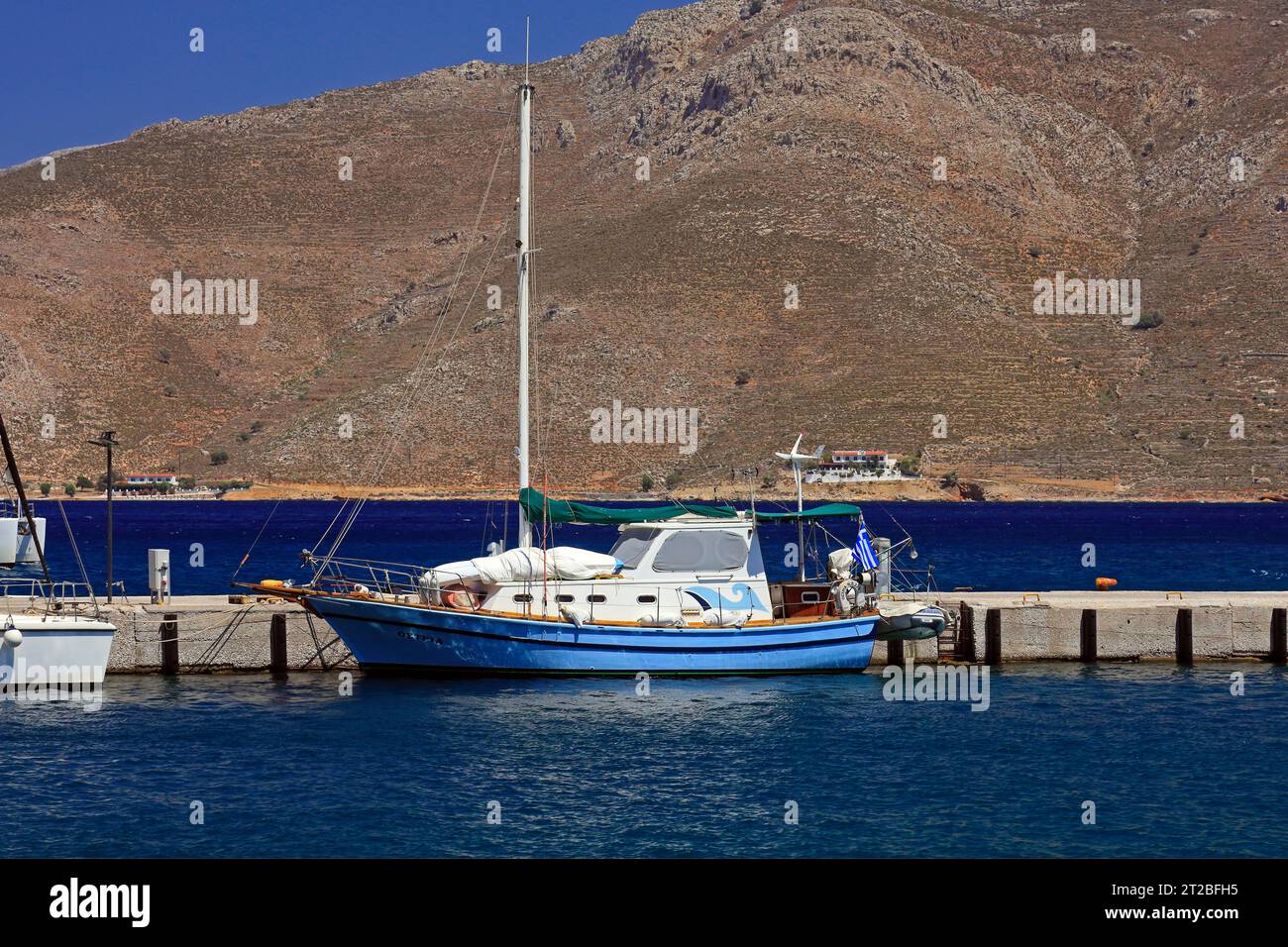 Blaues Segelboot, das im Hafen von Livadia mit den Bergen der hübschen Insel Tilos im Hintergrund vor Anker liegt. Dodekanese, Griechenland. Mai/Juni 2023 Stockfoto