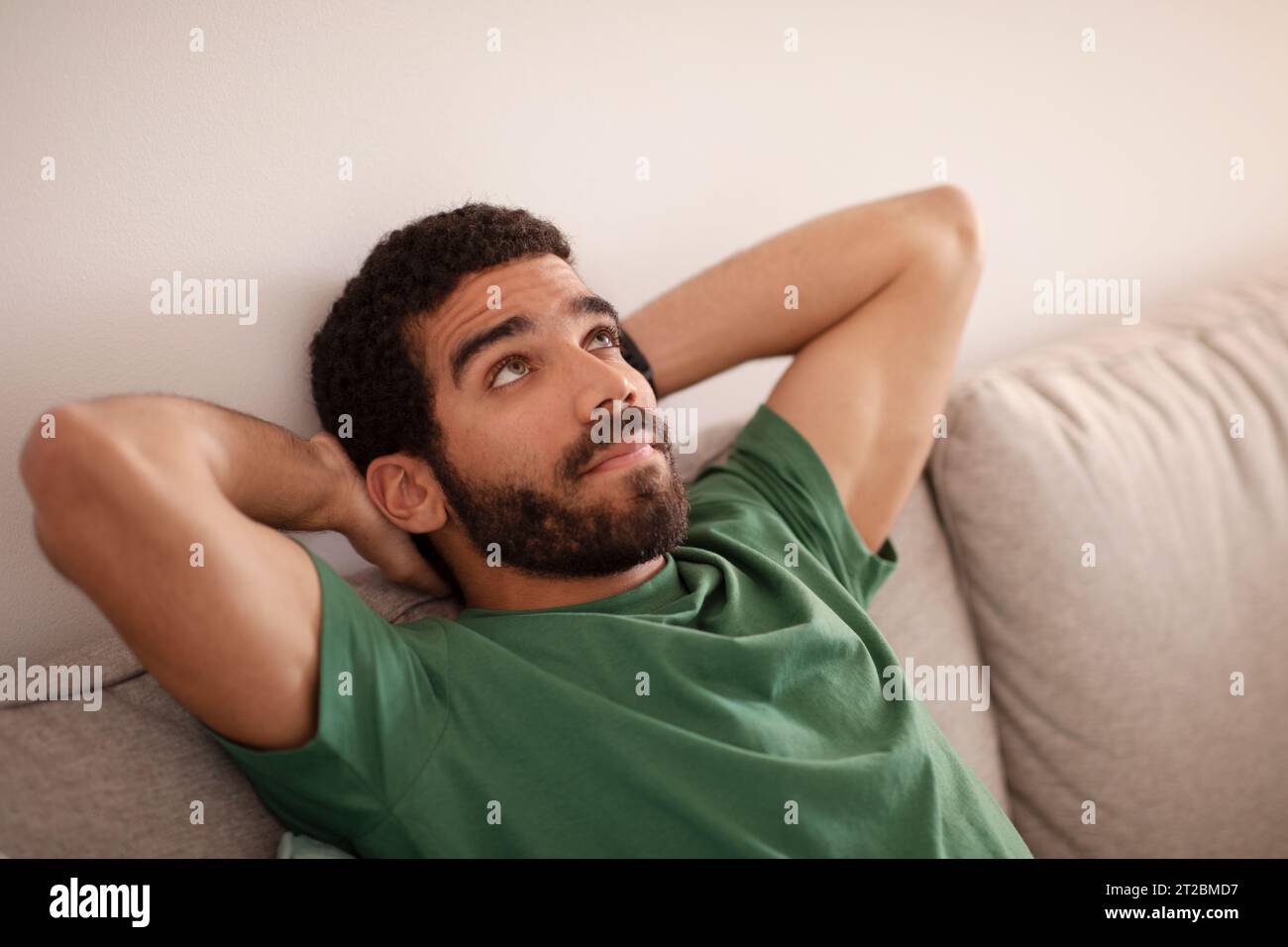 Nachdenklicher junger arabischer Mann mit Bart sitzt auf Sofa, ruht sich aus, schaut auf freien Raum, genießt Freizeit im Wohnzimmerinnenraum Stockfoto
