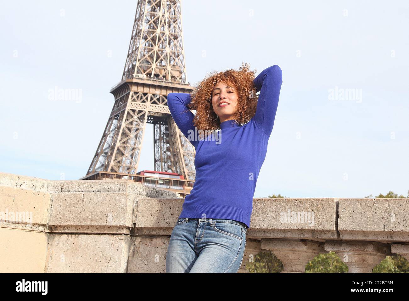 GLÜCKLICHE, LOCKIGE JUNGE FRAU IN BLAUEM PULLOVER, DIE SICH AM EIFFELTURM AMÜSIERT Stockfoto