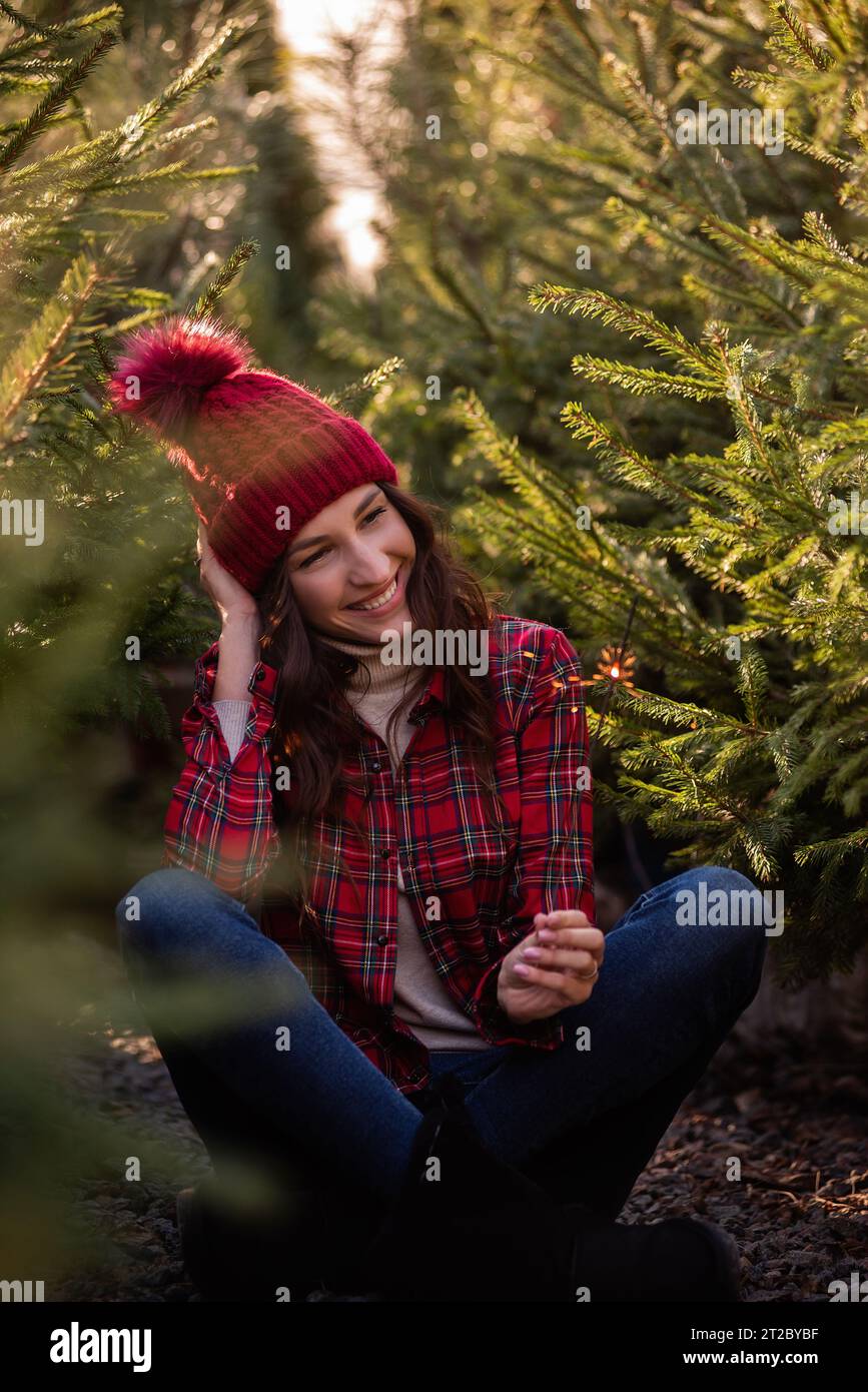 Nahporträt einer jungen Frau mit leuchtendem Funkeln in der Hand in rotem kariertem Hemd, Strickmütze zwischen grünen Kiefern. Lockiges Mädchen lacht, jubelt Stockfoto