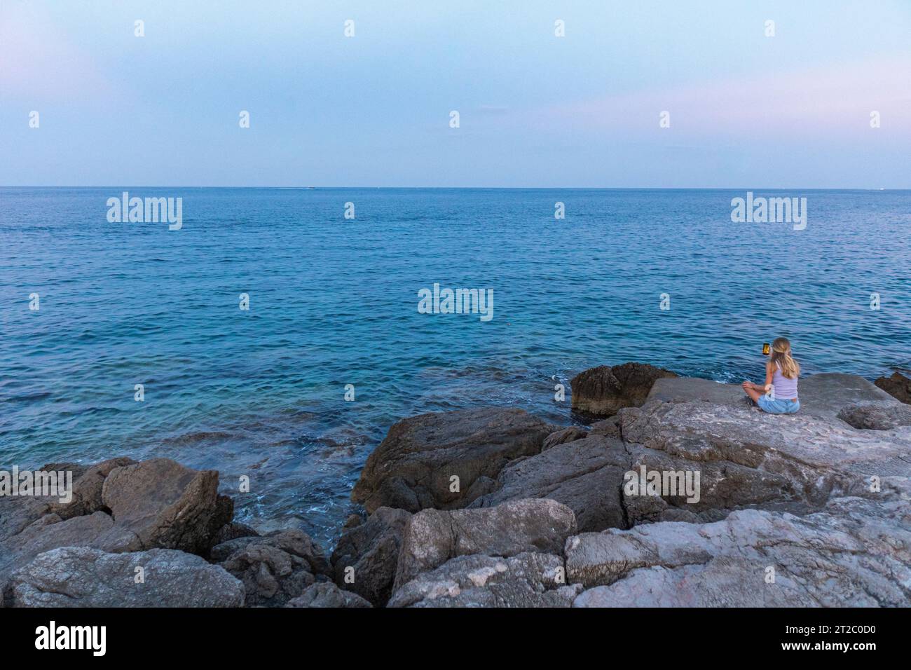 Ein blondes Mädchen, das auf den atemberaubenden Klippen von Cala Ratjada sitzt und das türkisfarbene Wasser des Mittelmeers auf Mallorca, Spanien, filmt Stockfoto