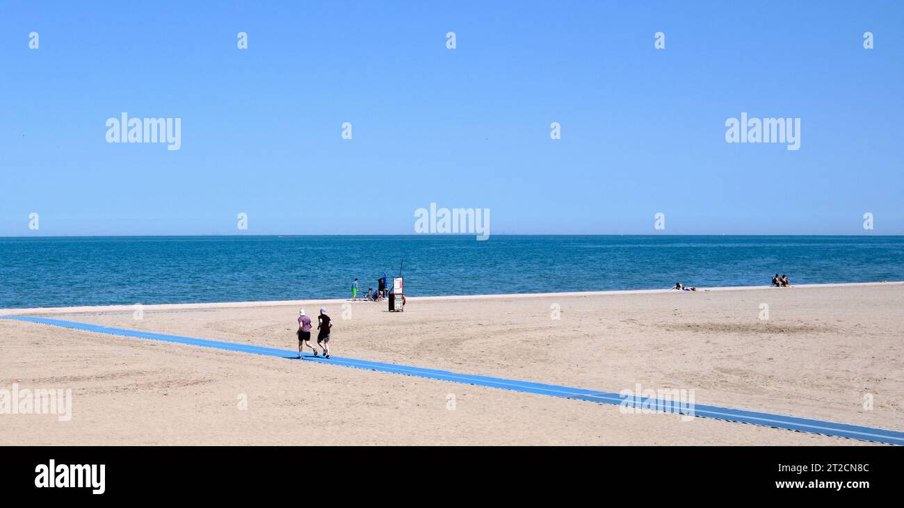 Zick-Zag-Muster von Boardwalk, Strand, Wasser und Horizont mit Joggern und Band aus blauem Fußweg über Sand. Minimalistische Fotografie. Stockfoto