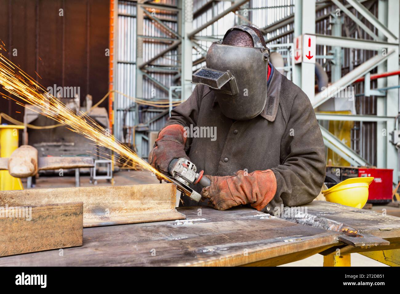 afrikanischer Mann in der Fabrik, der eine Maske trägt und mit einem Schleifer arbeitet, Funken fliegen Stockfoto