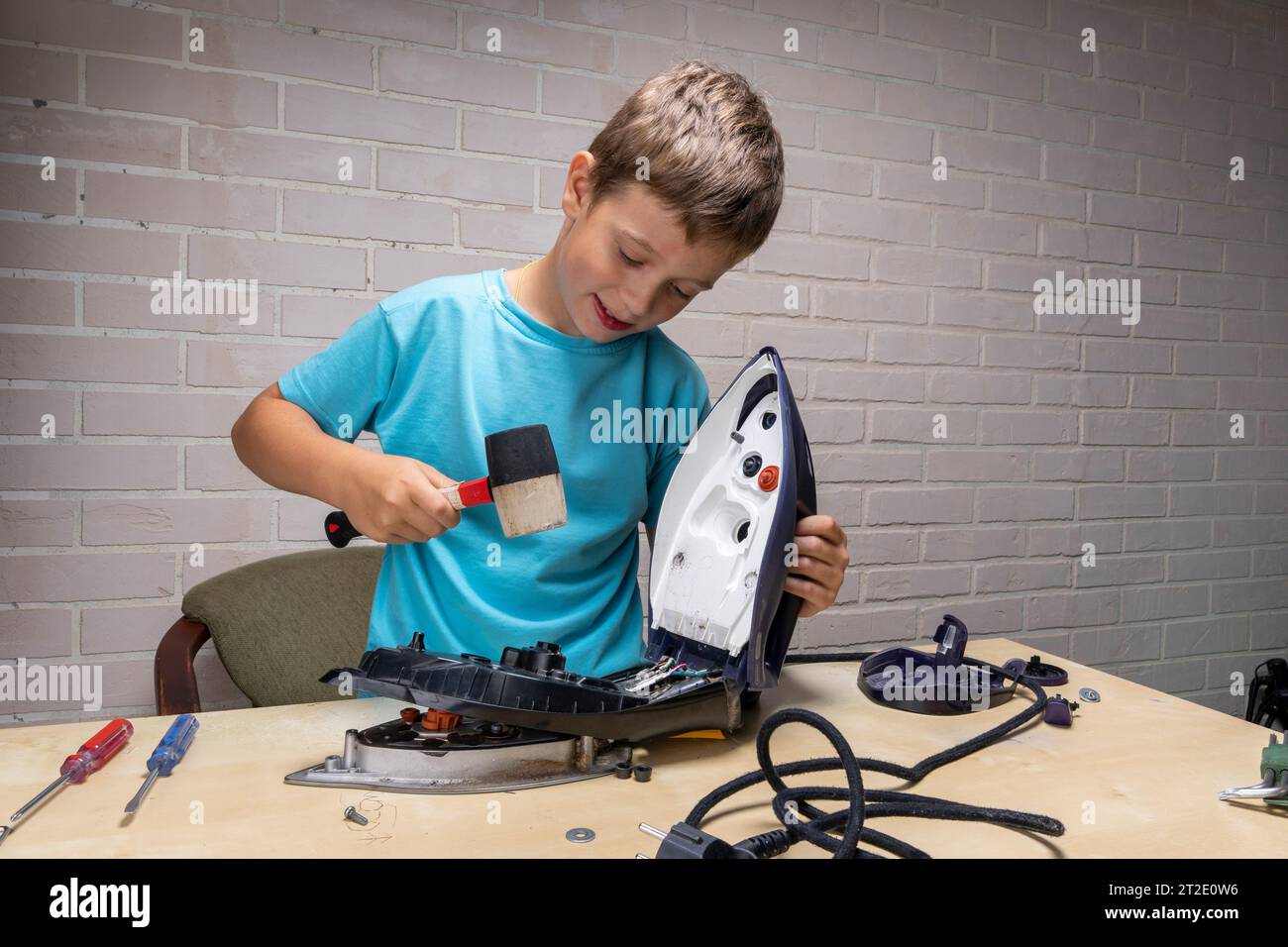 Junge hilft seiner Mutter. Ein lustiger Meister repariert ein Elektroeisen auf dem Tisch mit Werkzeugen. Zerlegtes Elektroeisen. Stockfoto