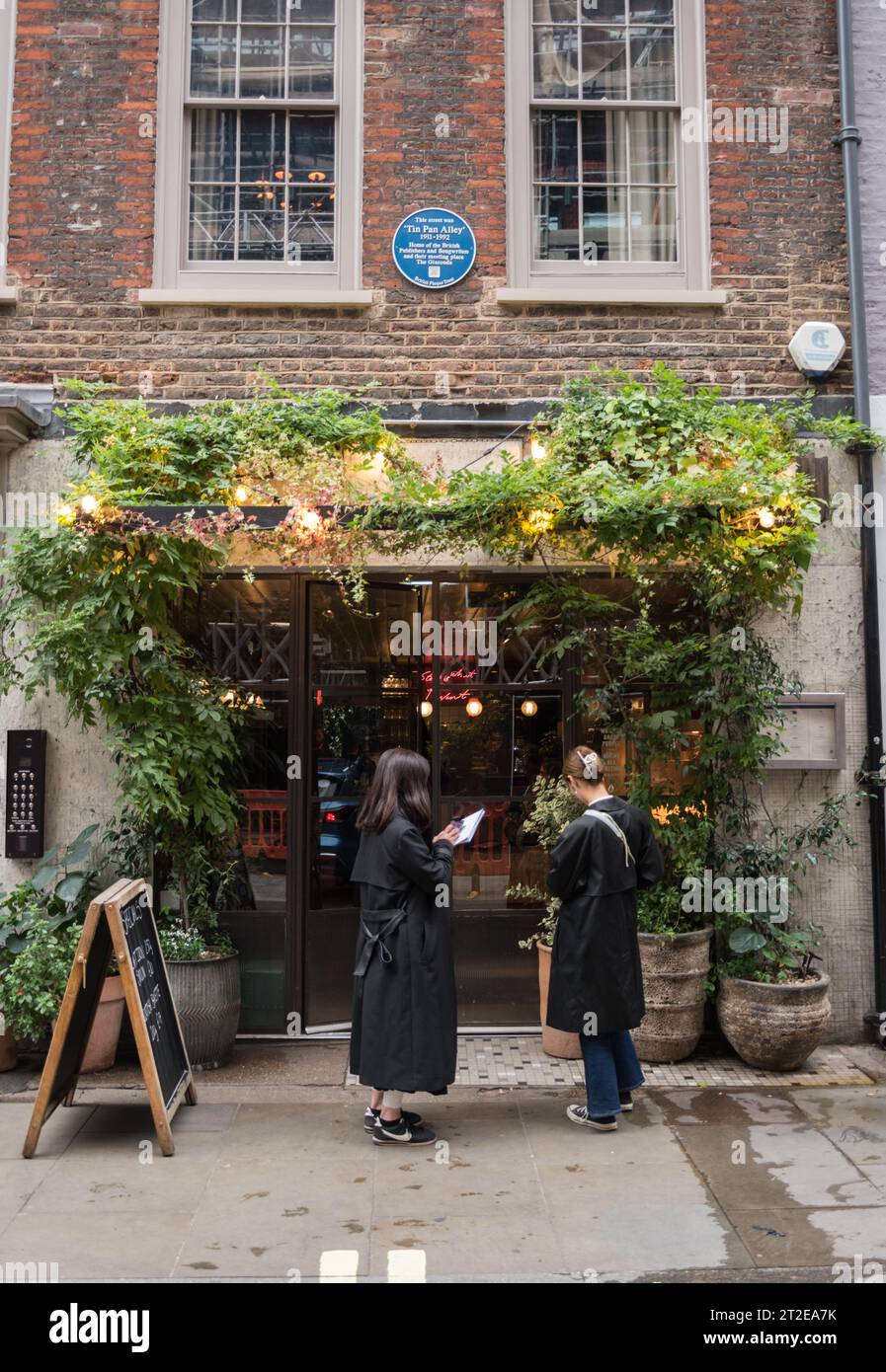 Gäste, die vor einem Restaurant unter einer blauen Tafel warten, die an die Lage der Tin Pan Alley in der Denmark Street, London, England, Großbritannien erinnert Stockfoto