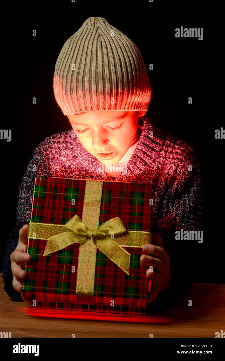 Ein Kind in warmer Kleidung und Wollmütze, das eine Geschenkbox öffnet, aus der ein leuchtendes Leuchten hervorgeht, mit einem überraschenden Gesichtsausdruck. Konz Stockfoto