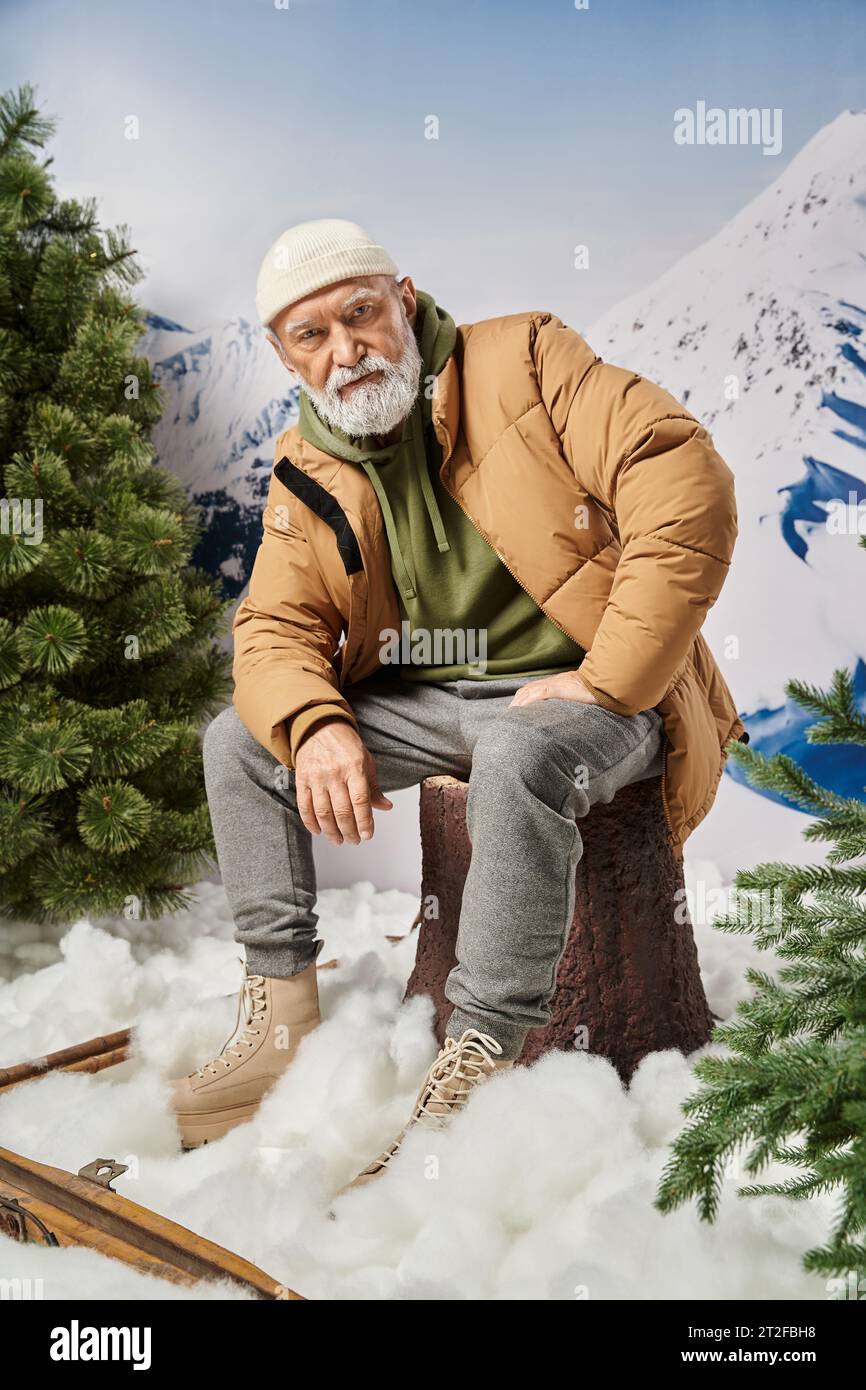 Ernster Mann mit weißem Bart in Jacke, der auf Baumstumpf sitzt und in die Kamera schaut, Winterkonzept Stockfoto