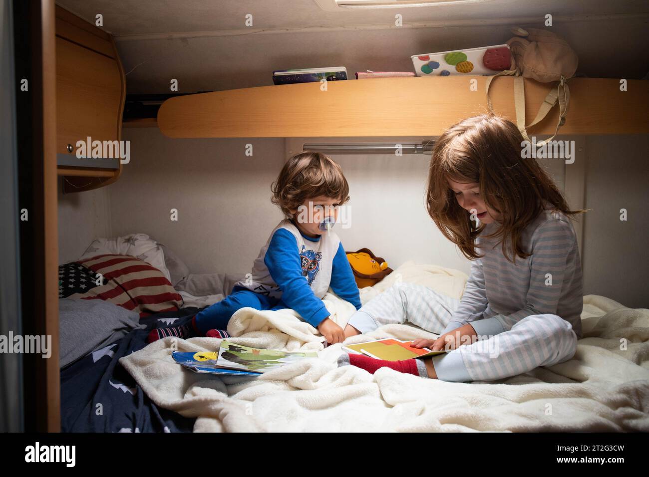 Zwei kaukasische Kinder, Bruder und Schwester, lesen Bücher im Pyjama auf einem Campervan-Bett während einer Autotour. Campingurlaub mit Kindern Stockfoto