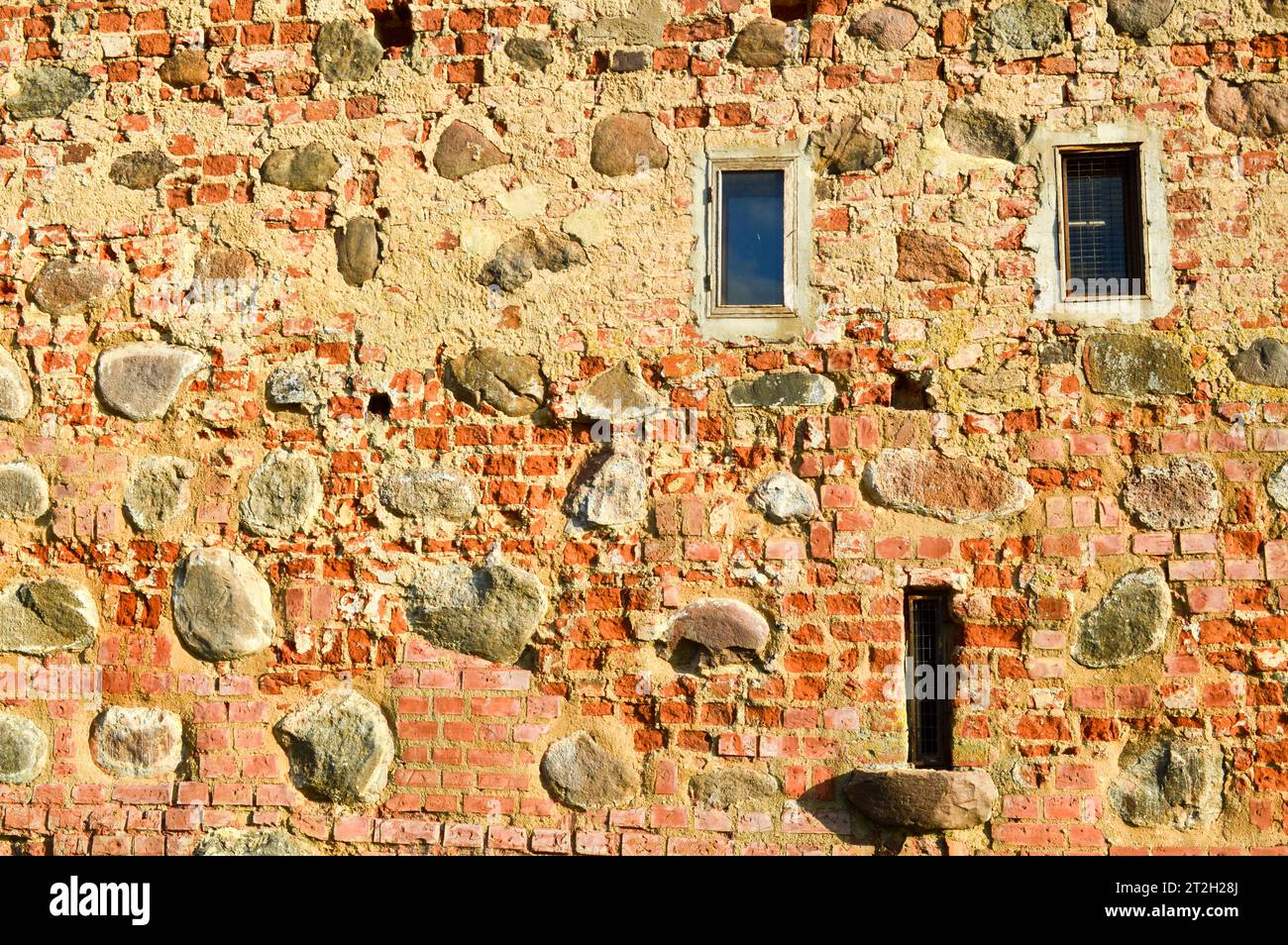 Textur mit kleinen, schmalen Fenstern auf einer alten alten Steinbruchwand aus rotem Backstein mit großen Felsblöcken. Der Hintergrund. Stockfoto