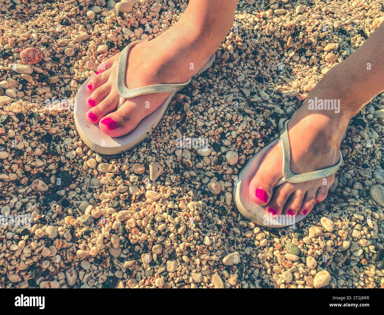 Ein Mädchen mit einer pinkfarbenen Pediküre an den Füßen spaziert in einem warmen Land in weißen Gummischuhen. Freizeitschuhe stehen auf dem Sand. Schieferplatten zum Gehen entlang der Stockfoto