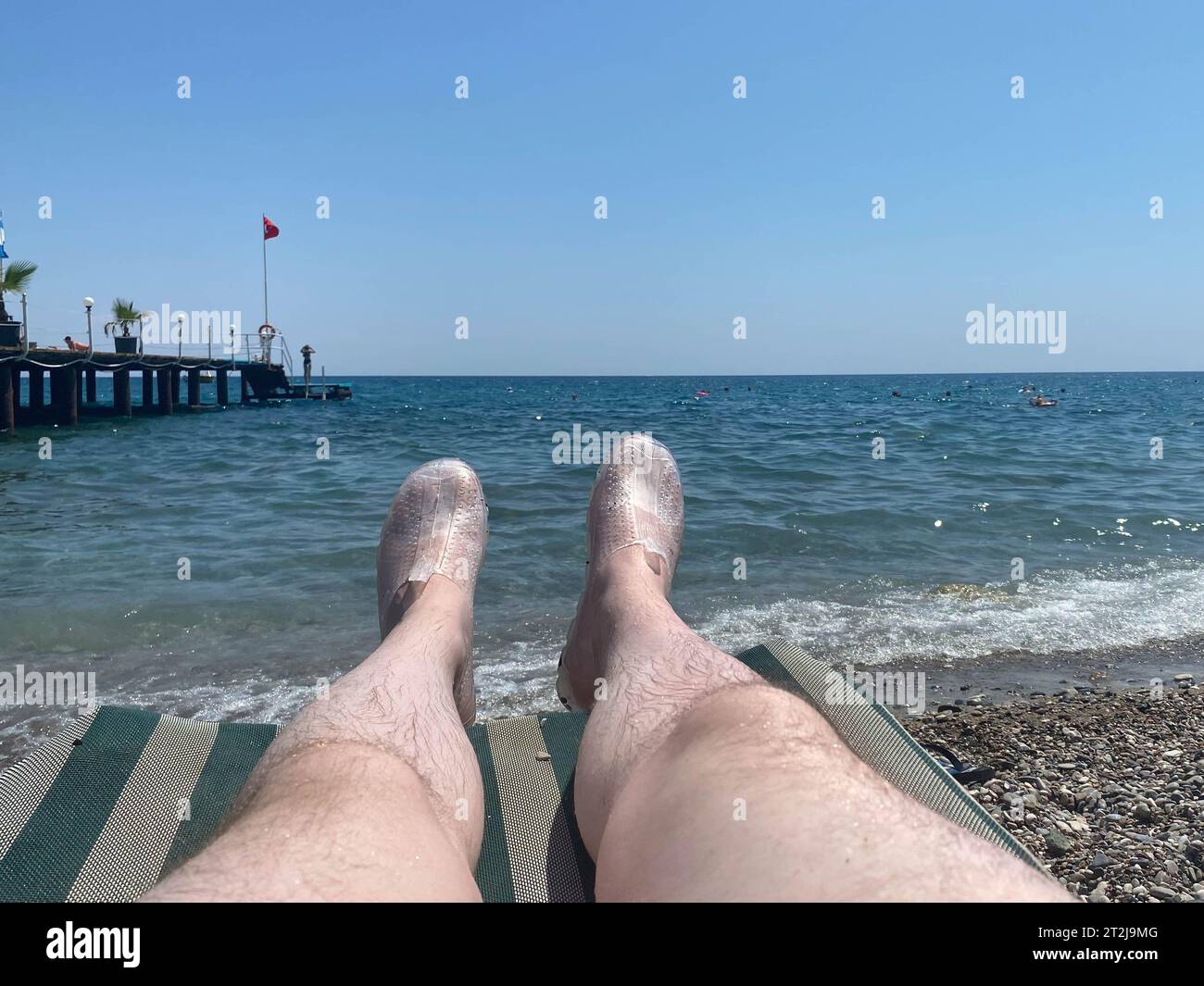 Junge Männer ruhen sich auf einer Strandliege in der Nähe des Schwimmbades aus, bei Sonnenuntergang mit blauem Wasser. Sommerurlaub. Männliche Beine liegen auf einer Sonnenbank am Meer. Stockfoto
