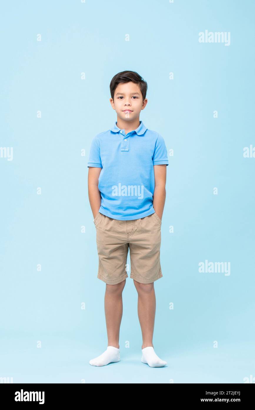 Selbstbewusstes Ganzkörperkind in legeren Kleidern und Socken, das Hände in Taschen hält und die Kamera vor blauem Hintergrund betrachtet Stockfoto
