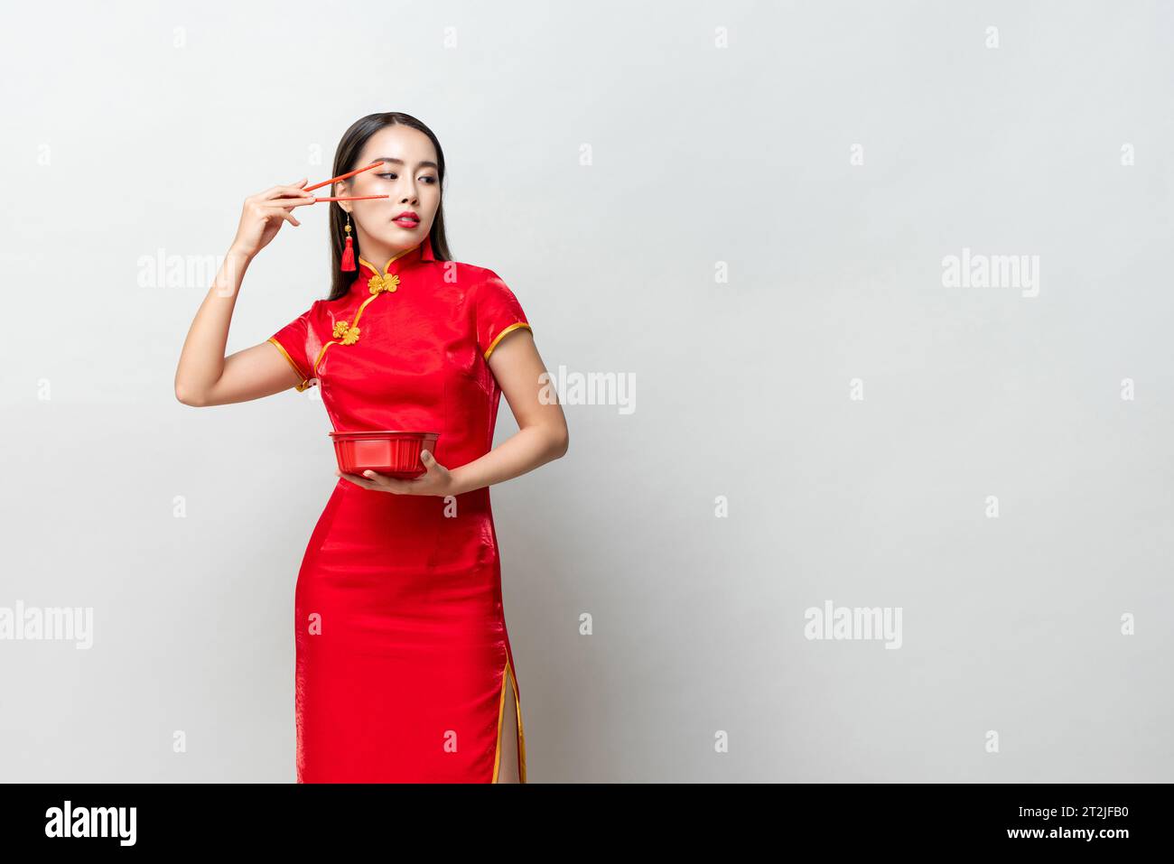 Asiatische Frau in traditionellem rotem Kleid, die den Arm mit Stäbchen hebt und auf den Kopierraum vor grauem Hintergrund blickt Stockfoto