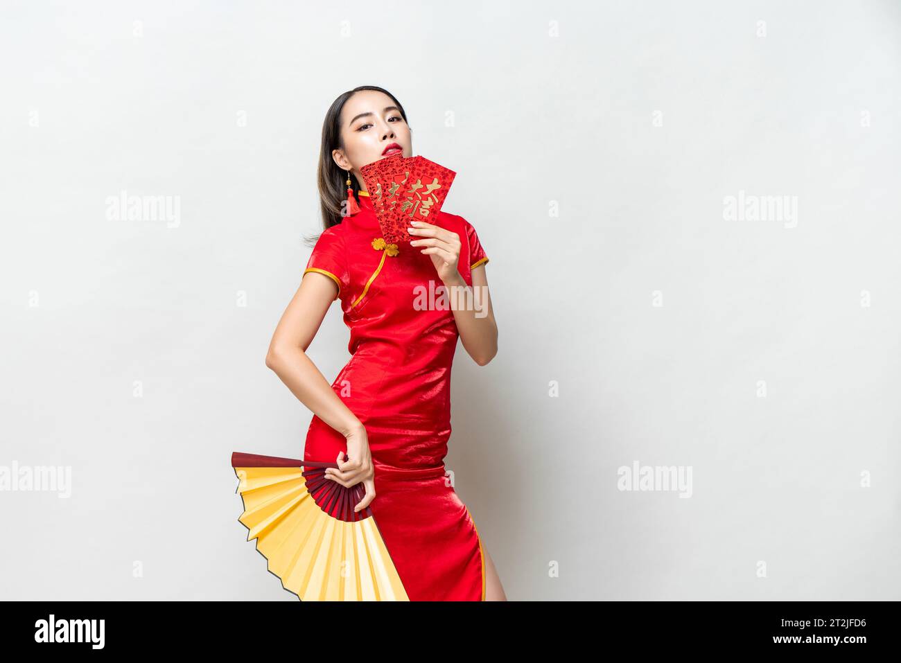 Asiatische Frau im orientalischen Cheongsam Kostüm, die rote Umschläge hält Ang Pow und Fan posiert auf hellgrauem Studiohintergrund für chinesische Neujahrskonzepte, Stockfoto