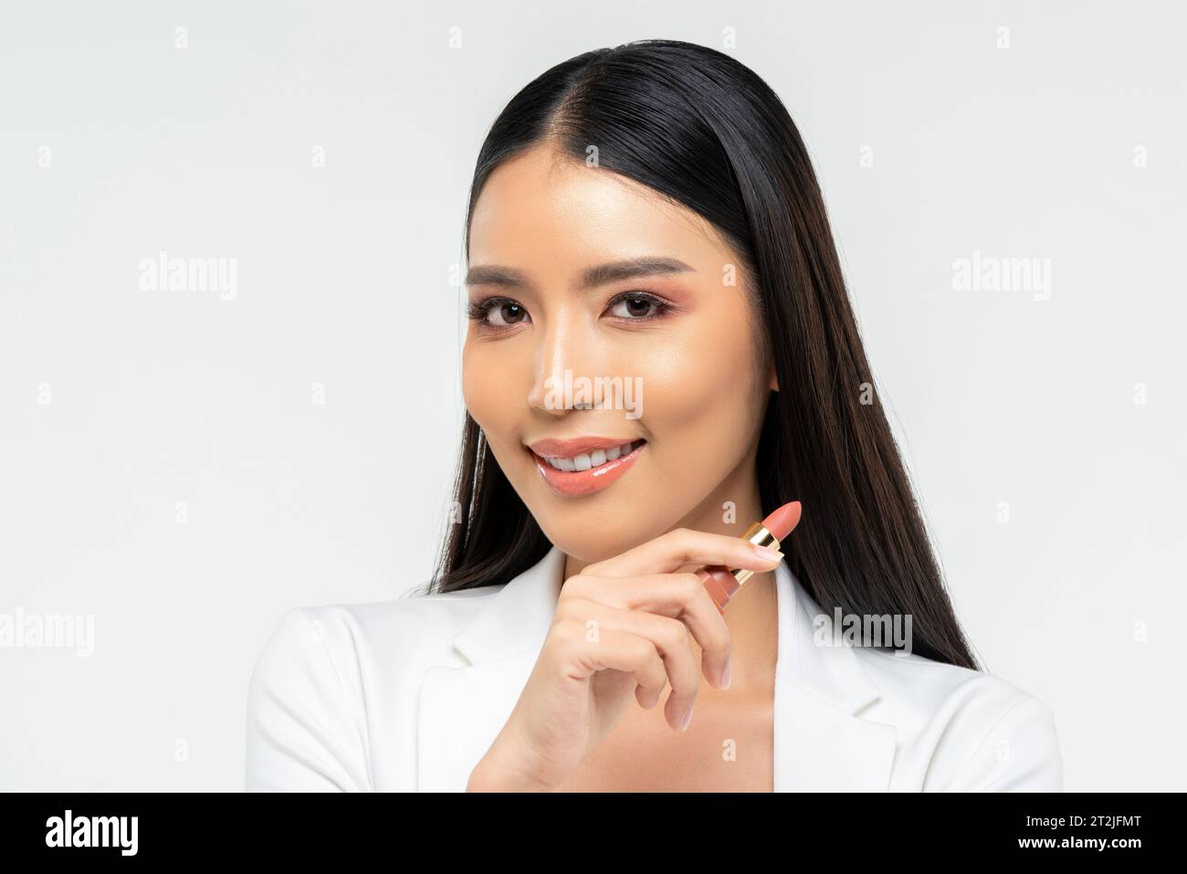 Freundliche junge asiatische Frau mit Lippenstift lächelt und während des Make-up-Auftrags in die Kamera blickt vor weißem Hintergrund Stockfoto