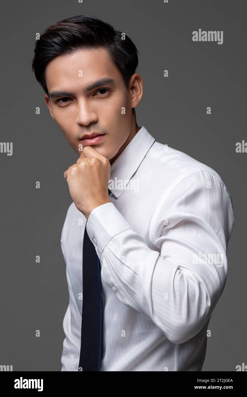 Seriöser asiatischer männlicher Unternehmer in weißem Hemd mit Krawatte berührendes Kinn auf grauem isoliertem Hintergrund im Lichtstudio Stockfoto