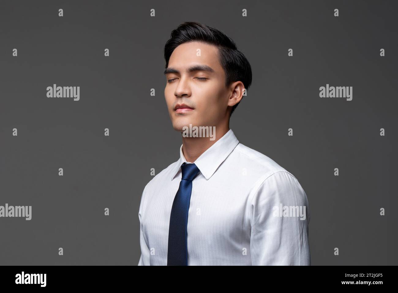 Schöner asiatischer Geschäftsmann in weißem Hemd mit Krawatte, der mit geschlossenen Augen auf grauem isoliertem Hintergrund im hellen Studio steht Stockfoto