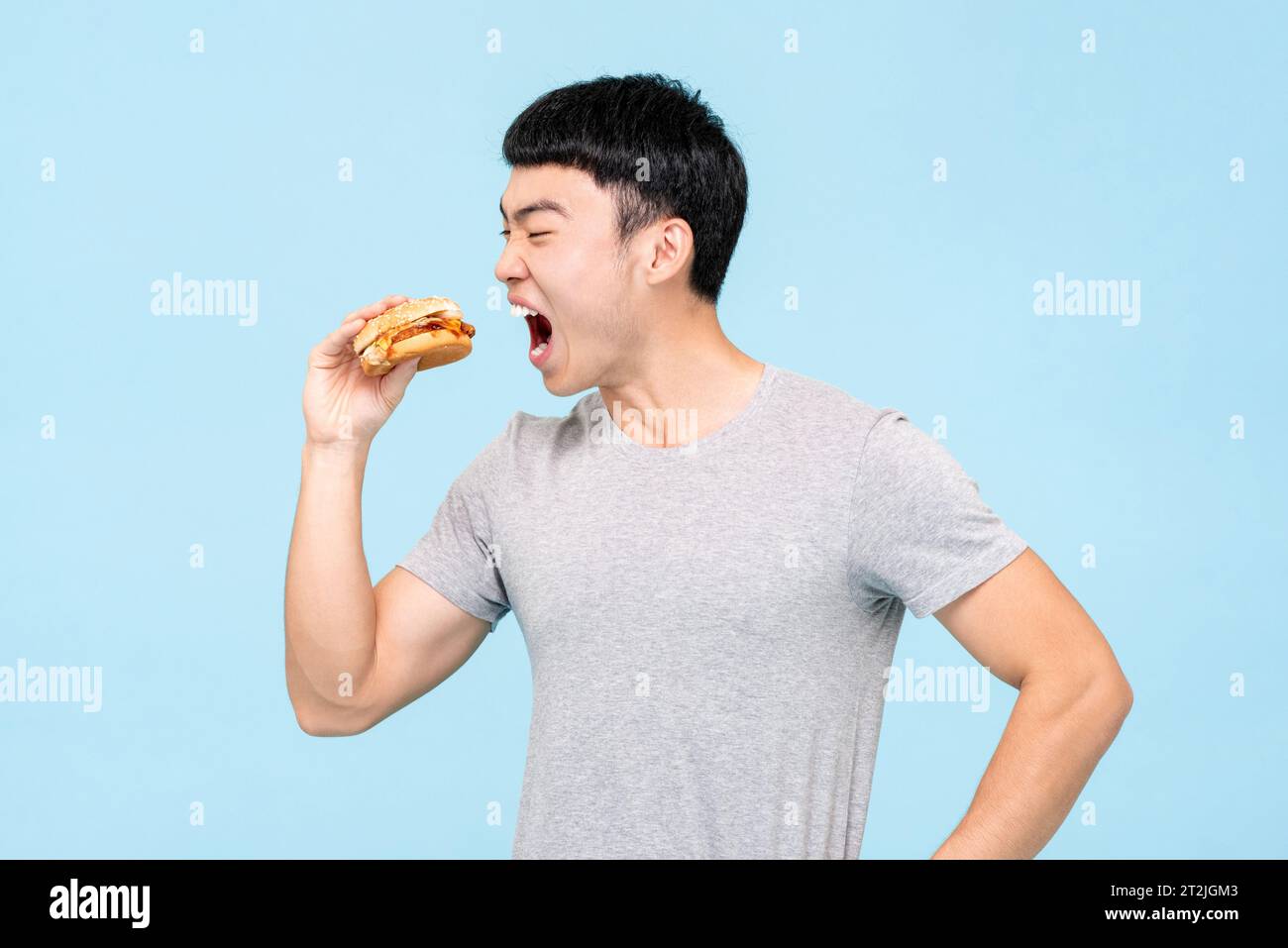 Hungriger asiatischer Mann in Casual Wear, der frischen ungesunden Burger isst, während er auf hellblauem isoliertem Hintergrund im Studio steht Stockfoto
